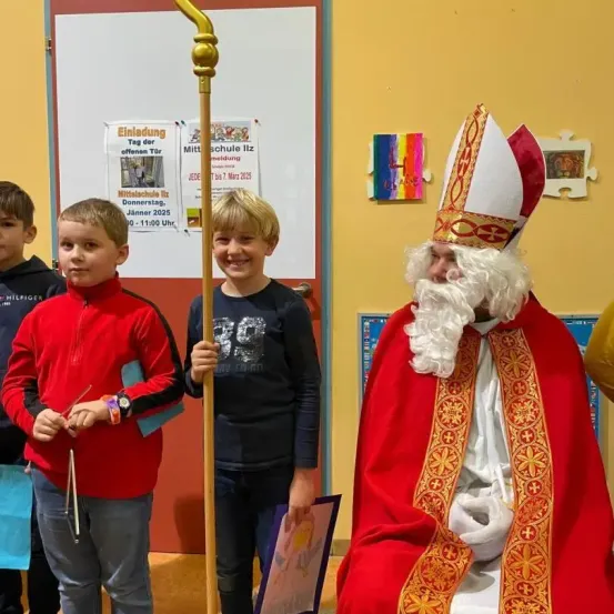 Drei Jungen und ein Mann, der als Papst verkleidet ist, stehen zusammen in einem Klassenzimmer. Die Jungen halten Stöcke, und einer trägt eine rote Jacke. Hinter ihnen steht eine Tafel mit einem Plakat, das einen Tag der offenen Tür an der Schule ankündigt.