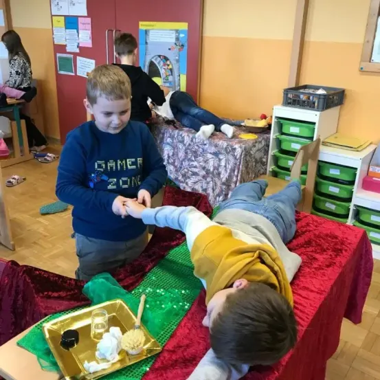 Zwei Jungen spielen in einem Klassenzimmer, einer liegt auf einem roten Tuch, während der andere seine Hand hält. Dahinter sitzt eine Frau auf einem Stuhl, andere stehen oder sitzen.