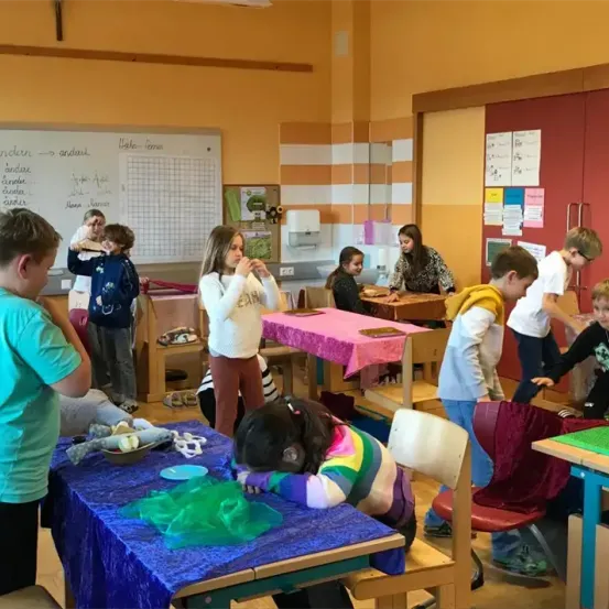 In einem Klassenzimmer sind Kinder mit verschiedenen Aktivitäten beschäftigt. Einige sitzen an Tischen mit Bastelarbeiten, während andere stehen und interagieren. Eine Tafel mit Text und Diagrammen ist an der Wand montiert.