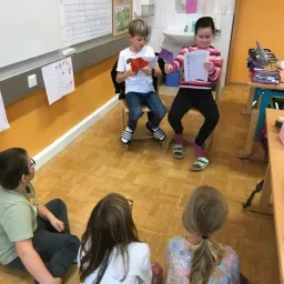 Mehrere Kinder sitzen in einem Klassenzimmer und lesen Papiere. Ein Junge sitzt auf einem Stuhl, während ein Mädchen ein Papier hält. Ein weiteres Mädchen sitzt auf dem Boden.