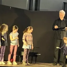 Ein älterer Mann steht mit einer Puppe und einem Mikrofon auf der Bühne, umgeben von fünf Kindern. Die Kinder beobachten ihn aufmerksam. Die Bühne ist schwach beleuchtet.