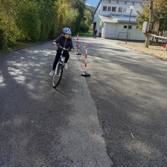 Ein junges Mädchen fährt mit einem Fahrrad auf einer leeren Straße, trägt einen Helm und Handschuhe. Es gibt Verkehrskegel entlang der Straße. In der Ferne ist ein Gebäude mit mehreren Fenstern zu sehen.
