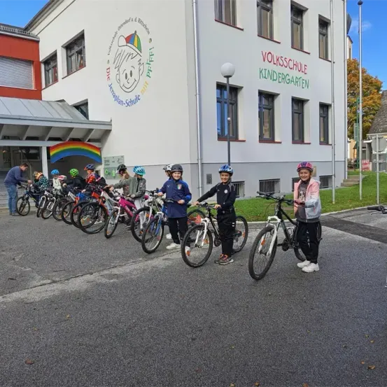 Kinder in Helmen und Fahrradkleidung stehen vor einem Gebäude, wahrscheinlich einer Schule, mit einem Regenbogen an der Wand.