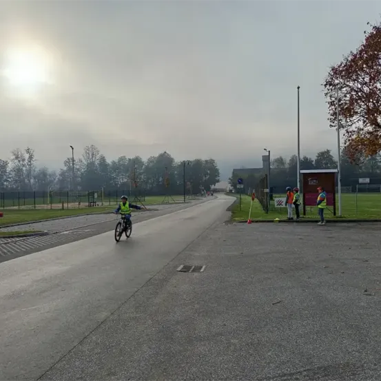 Ein Radfahrer in einer reflektierenden Weste fährt auf einer Straße mit einem Grasbereich und Bäumen. Drei Personen in Sicherheitswesten stehen in der Nähe eines Schildes.