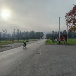 Ein Radfahrer in einer reflektierenden Weste fährt auf einer Straße mit einem Grasbereich und Bäumen. Drei Personen in Sicherheitswesten stehen in der Nähe eines Schildes.
