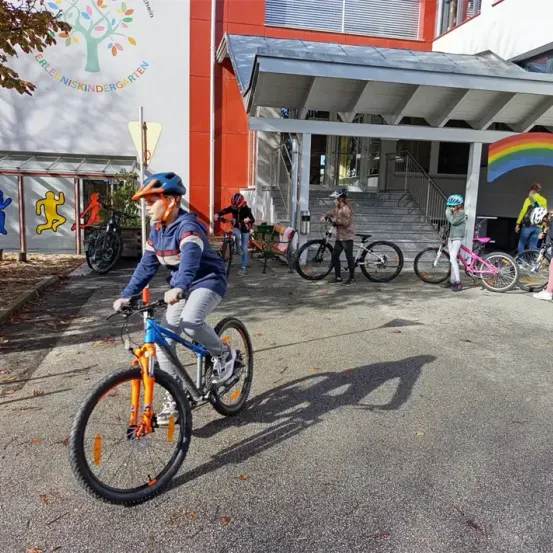 Mehrere Kinder fahren Fahrräder vor einem Kindergarten mit einem Regenbogenschild. Sie tragen Helme und einige haben Schuhe an.