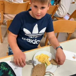 Ein Junge in einem marineblauen Adidas-Shirt sitzt an einem Tisch mit einem Teller Scrambled Eggs und Brot. Er hält eine Gabel und scheint zu essen.