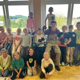 Eine Gruppe von Kindern und Jugendlichen lächelt und posiert für ein Foto in einem Raum mit Glasfenstern. Einige sitzen auf dem Boden, andere stehen hinter ihnen. Sie sind in verschiedenen Outfits gekleidet und lächeln fröhlich.