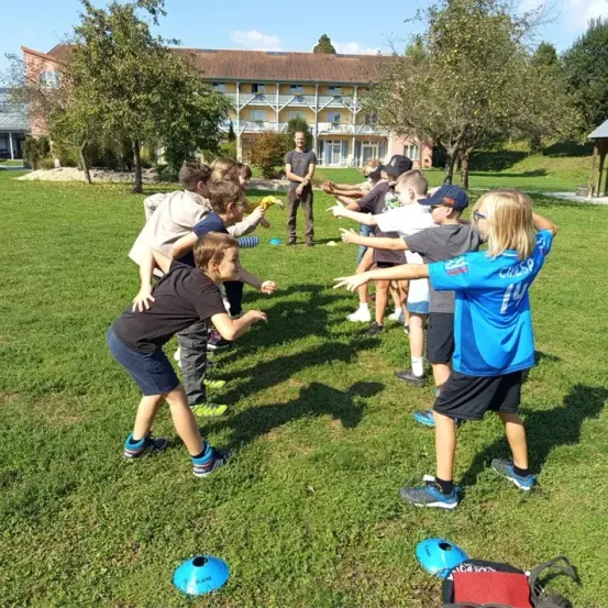 Kinder spielen auf einem Rasenfeld mit blauen Bällen und einem Trainer im Hintergrund. Das Gebäude mit Solarmodulen befindet sich dahinter.