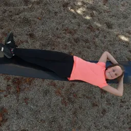 Ein Mädchen liegt auf einer Schaukel, trägt ein rosa Shirt und schwarze Hose und lächelt in den sonnigen Tag.