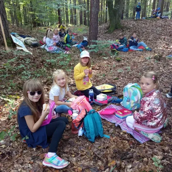 Eine Gruppe von Kindern macht ein Picknick im Wald. Sie sitzen auf dem Boden mit ihren Rucksäcken in der Nähe. Ein Mädchen isst ein Sandwich.