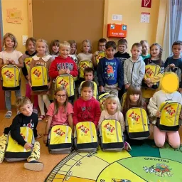 Eine Gruppe von Kindern mit Rucksäcken posiert für ein Foto in einem Klassenzimmer, stehend und kniend auf einem bunten Bodenteppich.