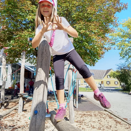 Ein Mädchen ist auf einem Klettergerüst in einem Park und posiert mit ihren Händen, die ein Herz formen. Hinter ihr befinden sich weitere Spielgeräte und Bäume. In der Ferne sind Häuser und ein geparktes Auto zu sehen.