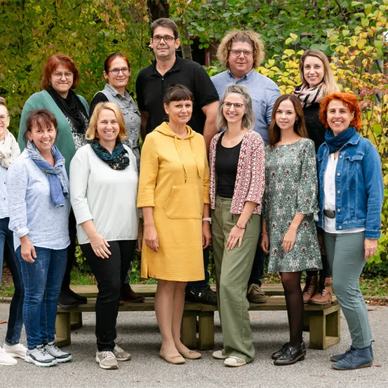Eine Gruppe von Menschen posiert für ein Foto im Freien, umgeben von grünem Laub, mit einer Holzbank im Vordergrund.