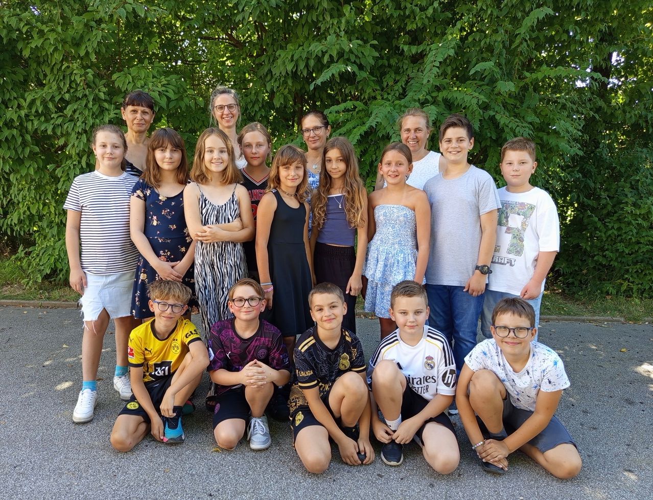Eine Gruppe junger Menschen und zwei Frauen posieren für ein Foto vor einem grünen Busch. Sie alle lächeln.