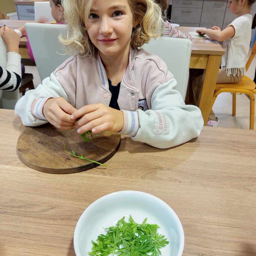 Ein kleines Mädchen sitzt an einem Tisch mit einem Holzschneidebrett und schneidet lächelnd Kräuter in eine Schüssel. Andere Kinder sitzen ebenfalls an Tischen im Hintergrund.