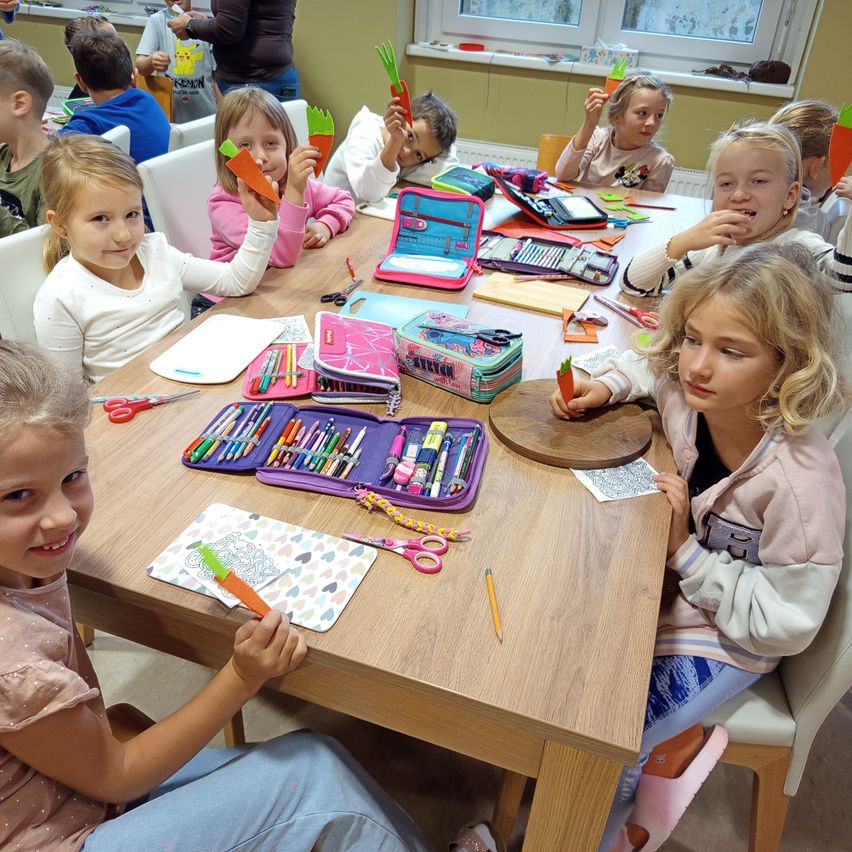 Mehrere junge Mädchen sind um einen Tisch mit verschiedenen Bastelutensilien versammelt. Jedes Mädchen hält eine karottenförmige Bastelei in der Hand, und sie scheinen sich in einem Klassenzimmer zu befinden.