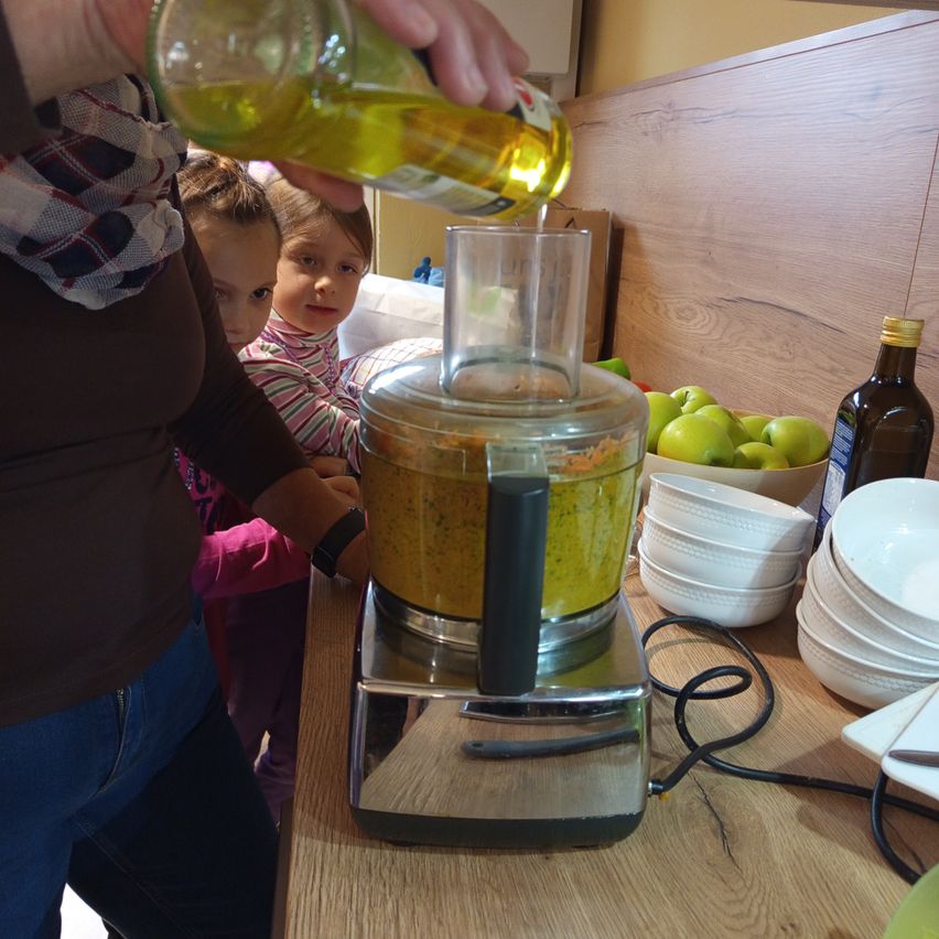 Eine Person gießt Öl in einen Foodprozessor, während zwei Kinder zusehen. Der Tresen hat Teller, Äpfel und eine Ölflasche.