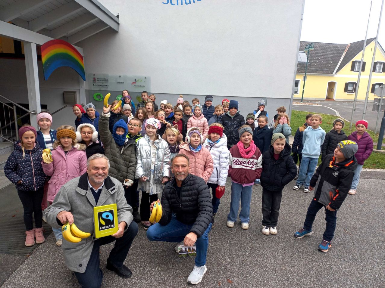 Eine Gruppe von Kindern und Erwachsenen posiert für ein Foto vor einem Schulgebäude, wobei zwei Männer Bananen halten und lächeln.