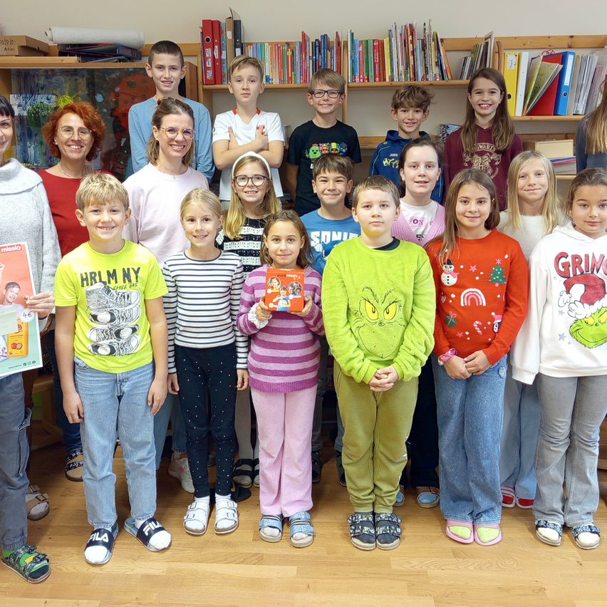 Eine Gruppe von Kindern und Erwachsenen posiert für ein Foto in einer Bibliothek, hält Bücher in den Händen und trägt festliche Pullover.