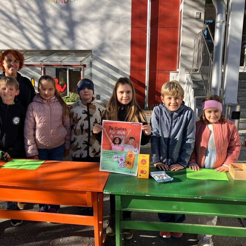 Eine Gruppe von Kindern und einem Erwachsenen stehen hinter Tischen im Freien und halten ein Poster und Snacks. Die Kinder lächeln und im Hintergrund befindet sich ein Gebäude mit einer Treppe.