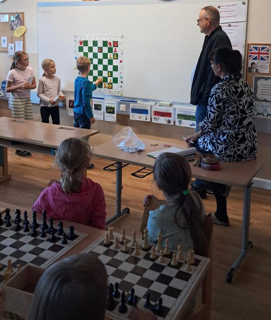 Ein Klassenzimmer mit Schülern, die Schach spielen. Ein Lehrer beobachtet. Schachfiguren sind auf den Brettern und einige Kinder stehen in der Nähe einer Tafel mit einem Schachdiagramm.