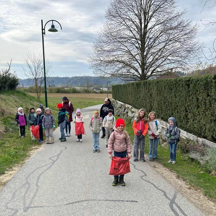 A group of children and adults are walking on a path, holding red bags. They are wearing winter clothes and some have hats. A street light and a tree are on the left side.