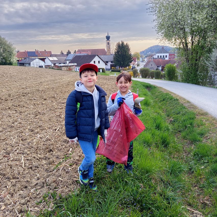 Two boys are smiling and collecting trash in a field, with houses and a tower in the background.