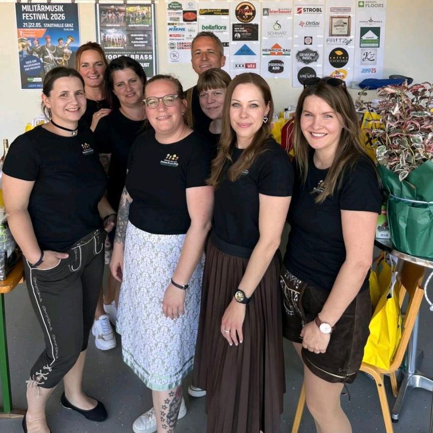 A group of smiling women in black shirts stand together in a room with promotional posters. Two women wear skirts, and one has a tattoo on her leg.