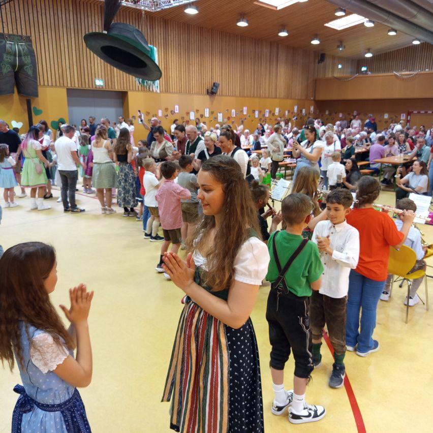 Inside a large hall, a diverse crowd, including children and adults, are gathered. A girl in a colorful dress and two children in traditional attire are clapping. The atmosphere is festive with wooden walls and hanging decorations.