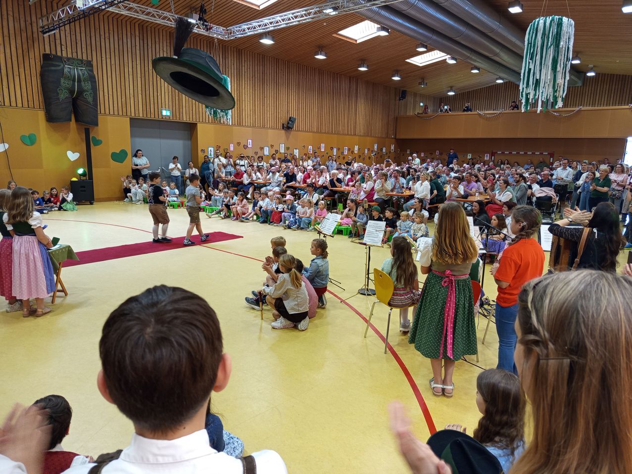 A large audience is gathered indoors, watching children perform on a red carpet. Some are seated, while others stand. A woman with a green dress and a ribbon around her waist stands before a microphone.