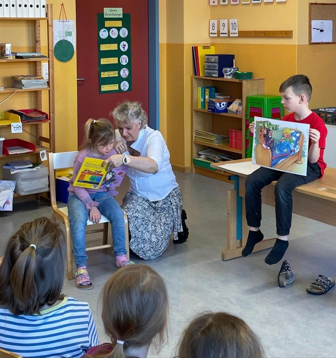 Eine Frau liest einem Kind in einem Klassenzimmer ein Buch vor. Andere Kinder sitzen um sie herum. Es gibt Regale mit Büchern und Nummern an der Wand.