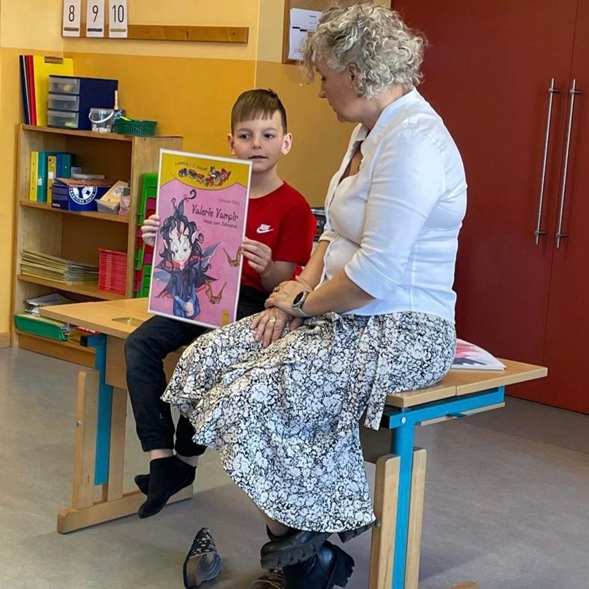 Eine Frau liest einem Jungen vor, der an einem Schreibtisch in einem Klassenzimmer sitzt. Der Junge hält ein Buch mit dem Titel Valerie Vampir.