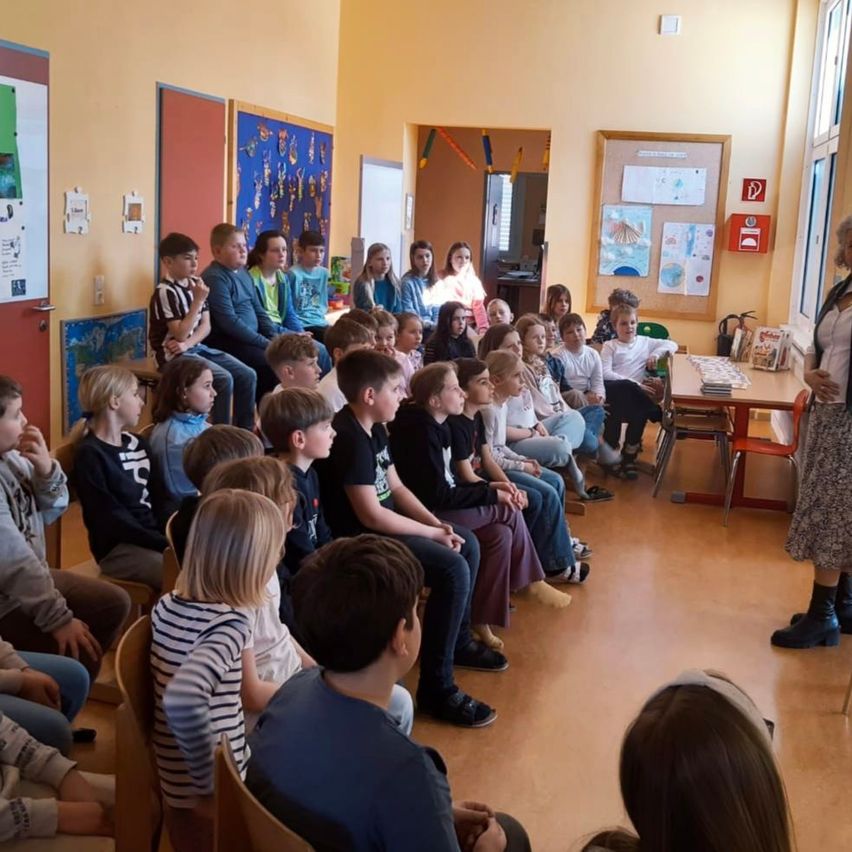 Ein Klassenzimmer voller Kinder, die einer Frau beim Sprechen aufmerksam zuhören, mit verschiedenen Kunstwerken und einem Tisch mit Büchern.