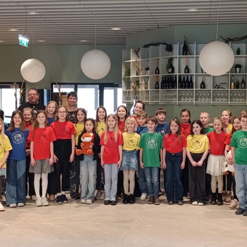 Eine Gruppe junger Schüler und Erwachsene posiert für ein Foto in einem Raum mit Regalen, Flaschen und Luftballons. Die Kinder tragen passende T-Shirts.
