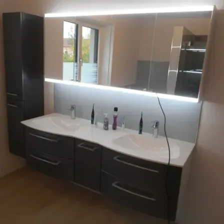 Ein modernes Badezimmer mit Doppelwaschbecken, großem Spiegel mit Lichtleiste und hoher Kommode. Der Boden ist aus Holz.