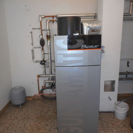Ein Raum mit einer grauen Biostar-Heizungsanlage an einer weißen Wand, Kupferrohren und einem Eimer in der Nähe.