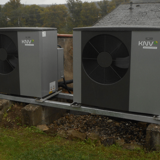 Zwei graue KNV-Klimageräte sind auf einem Metallrahmen montiert und auf einem Rasen mit Steinen und Kies positioniert.
