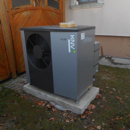 Eine graue Klimaanlage steht draußen auf einem Betonblock mit umherliegenden Blättern. Ein gelbes Objekt befindet sich oben auf der Einheit.