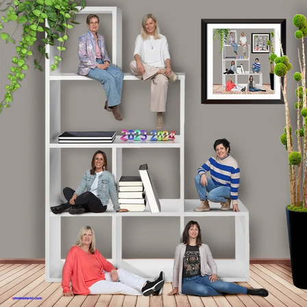 Eine Gruppe von Frauen posiert für ein Foto vor einem weißen Regal mit Büchern und einem 2023-2024-Schild. Auf beiden Seiten des Regals stehen zwei große Topfpflanzen.