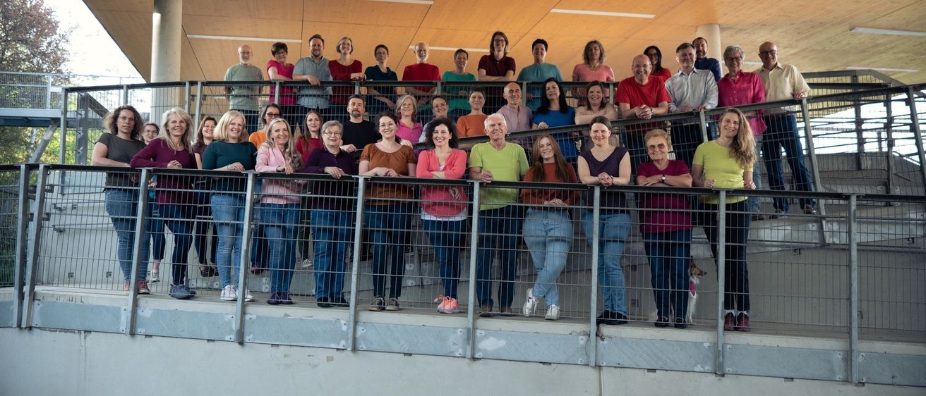 Eine Gruppe von Menschen in verschiedenen farbigen T-Shirts steht in einer Reihe auf einer Terrasse. Die Terrasse hat Metallgeländer. Die Menschen lächeln und scheinen für ein Foto zu posieren. Hinter ihnen ist eine Holzdecke und eine weiße Säule. Die Menschen tragen T-Shirts, Hosen und Schuhe in verschiedenen Farben.