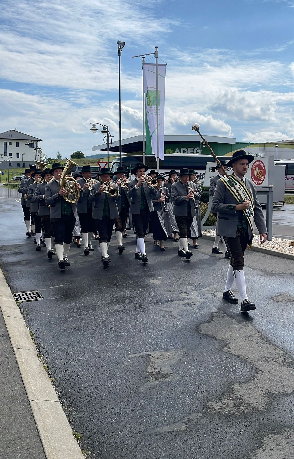 Eine Blaskapelle, in traditioneller Kleidung gekleidet, spielt Instrumente, während sie auf einer nassen Straße geht, mit Gebäuden und Straßenlaternen im Hintergrund.
