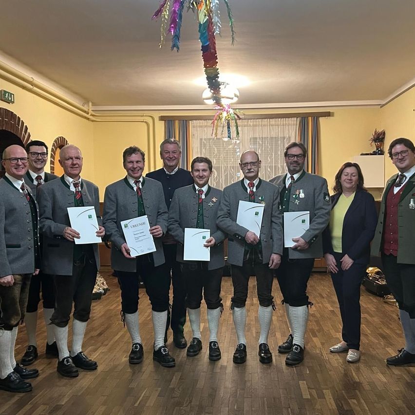A group of individuals in traditional German attire poses for a photo, holding certificates. The setting appears to be a celebratory event, with decorations and a festive atmosphere.
