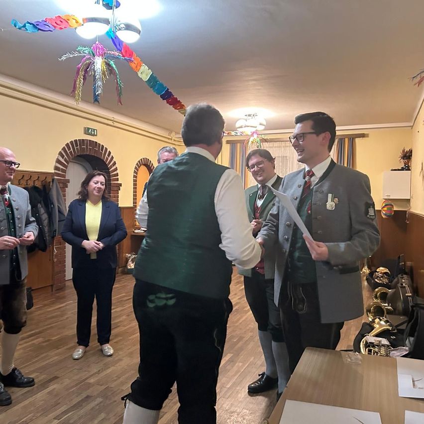 A group of people are standing in a room. One man is holding a paper, while another is shaking his hand. They are wearing traditional costumes. The room has wooden floors, a brick wall, and decorations. There is a desk with papers and musical instruments.