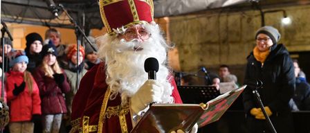 Ein Mann, der als Weihnachtsmann verkleidet ist, hält ein Mikrofon und liest aus einem Buch. Dahinter sieht man eine Menge, die zuschaut, und Musikinstrumente.