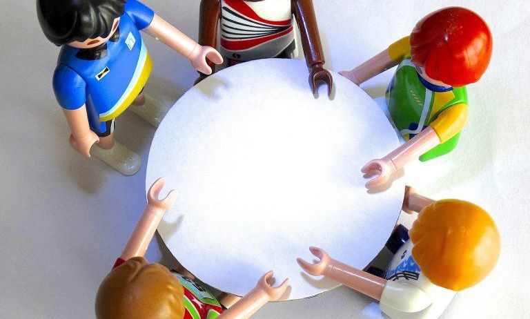 Fünf Spielzeugfiguren stehen um einen weißen runden Tisch herum, wobei jede einen Teil des Randes hält. Eine Figur trägt ein blaues Shirt und hat langes schwarzes Haar.