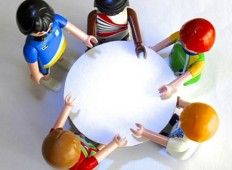 Fünf Spielzeugfiguren stehen um einen weißen runden Tisch herum, wobei jede einen Teil des Randes hält. Eine Figur trägt ein blaues Shirt und hat langes schwarzes Haar.