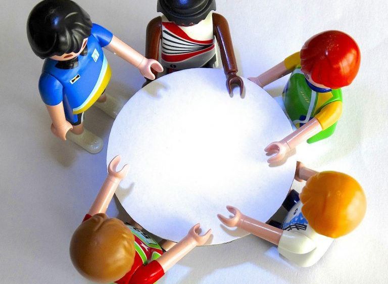 Fünf Spielzeugfiguren stehen um einen weißen runden Tisch herum, wobei jede einen Teil des Randes hält. Eine Figur trägt ein blaues Shirt und hat langes schwarzes Haar.