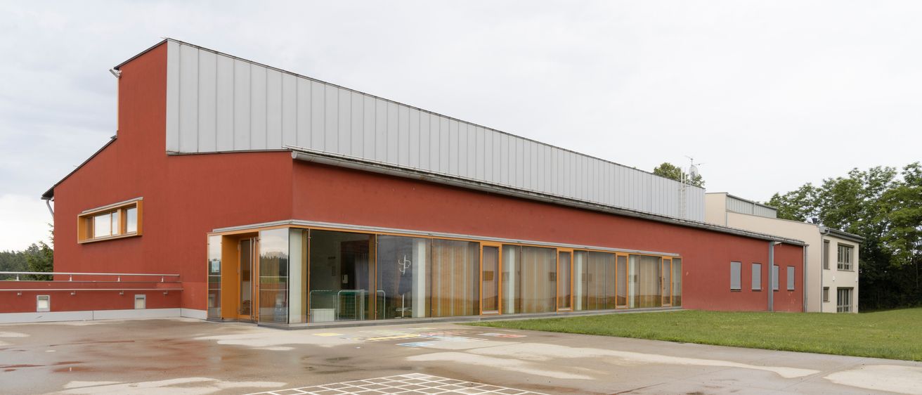 Ein rotes Gebäude mit grauem Dach und großen Glasfenstern ist zu sehen. Das Gebäude hat orangefarbene Einfassungen um die Fenster. Ein Spielplatz befindet sich vor dem Gebäude.