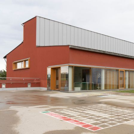 Ein großes rotes Gebäude mit einem weißen Dach und Glasfenstern steht draußen. Vor dem Gebäude befindet sich ein Spielplatz.